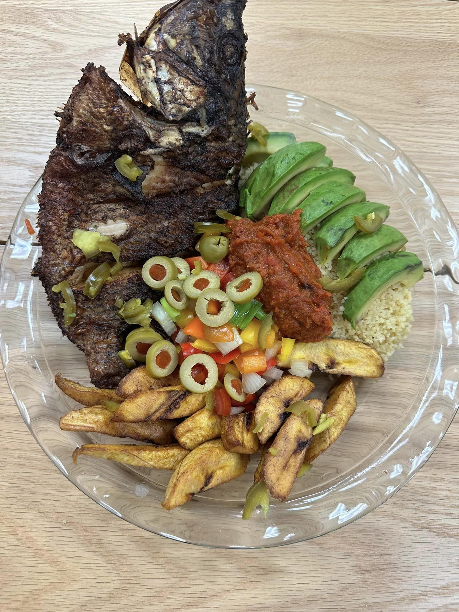 A clear plate contains a grilled fish, sliced avocado, fried plantains, couscous, a vegetable mix with olives, and a serving of red sauce.