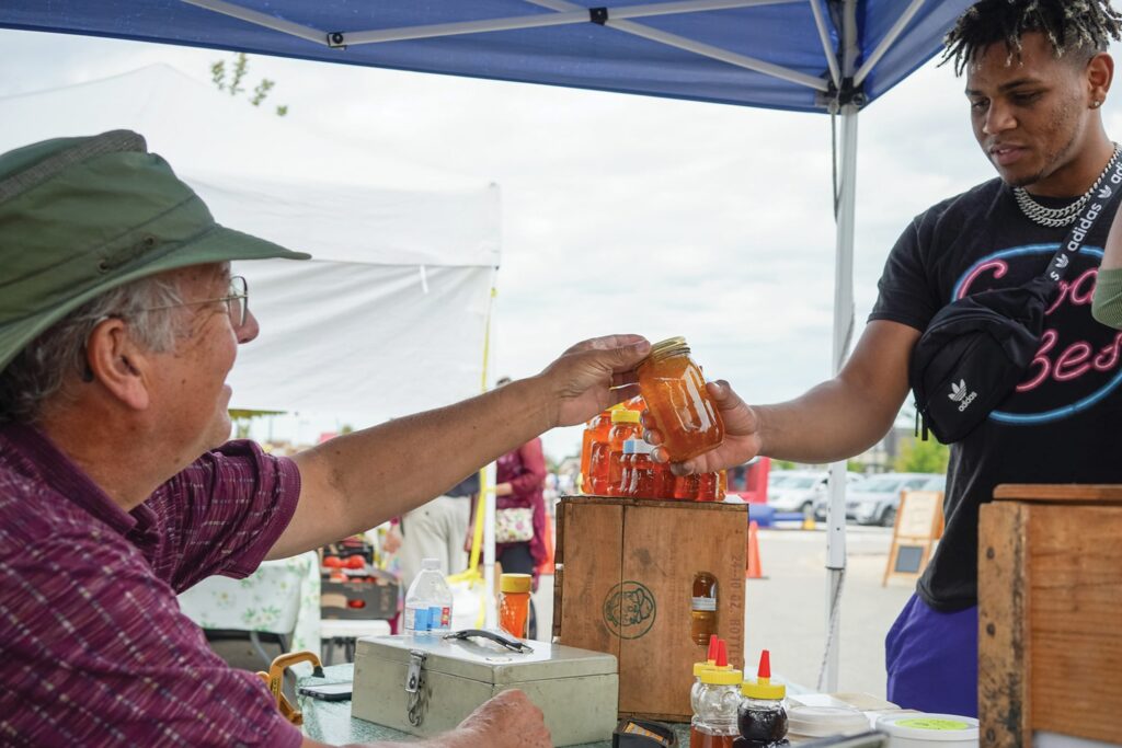 A man wearing a green hat and plaid shirt hands a jar of honey to a customer under a blue canopy at an outdoor market.