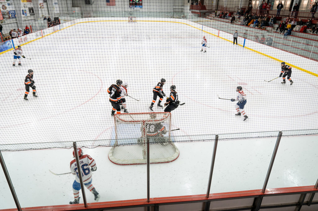 A hockey game in progress at an indoor ice rink, with players in black and yellow jerseys spread across the rink and spectators visible in the stands.