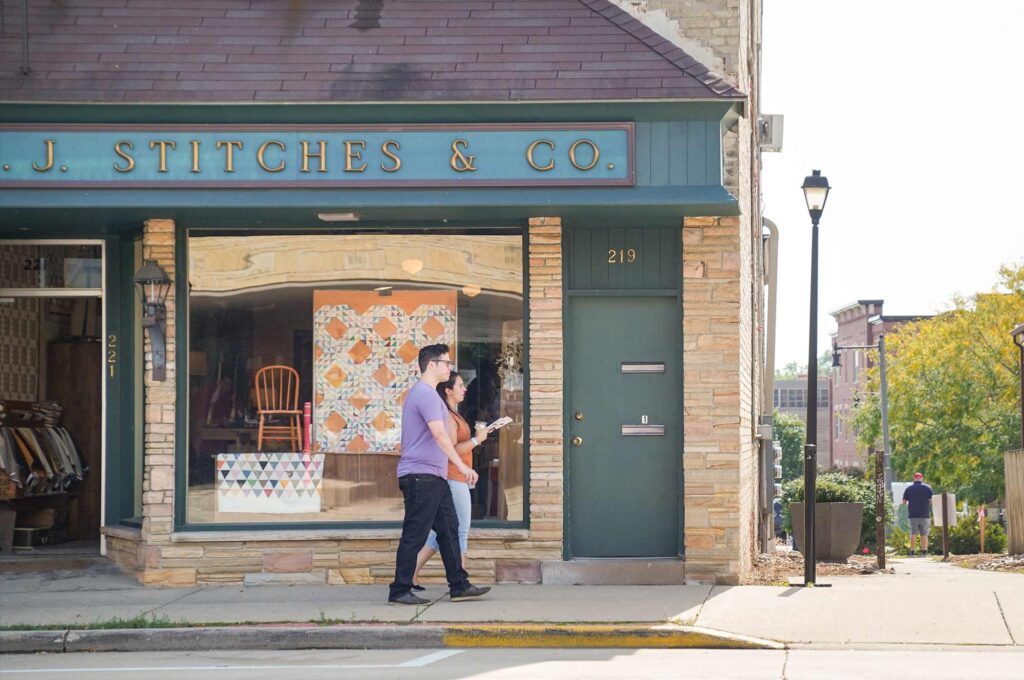 A man and woman walk past a storefront named "J. Stitches & Co." on a sunny day. The store has a large display window featuring a quilt and a wooden chair.