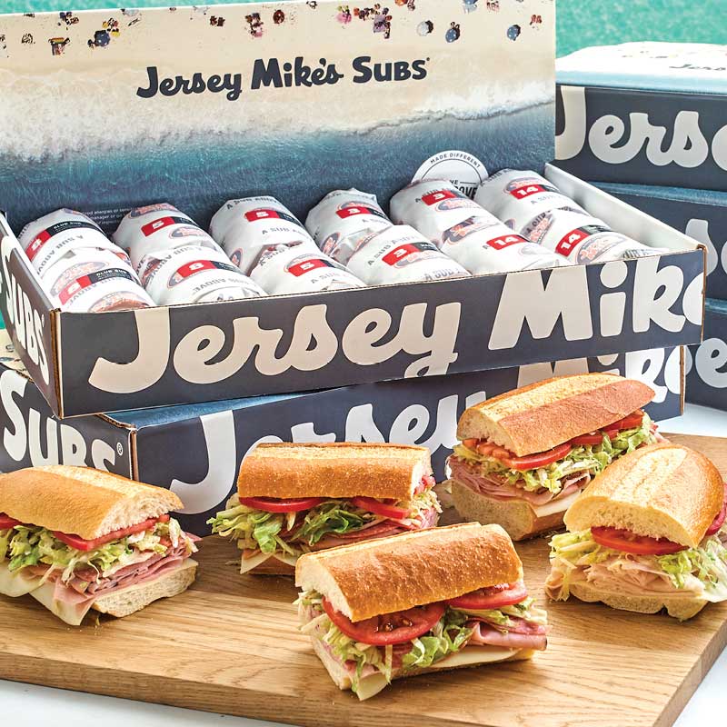 Assorted subs on a wooden board in front of Jersey Mike's boxes filled with canned drinks.