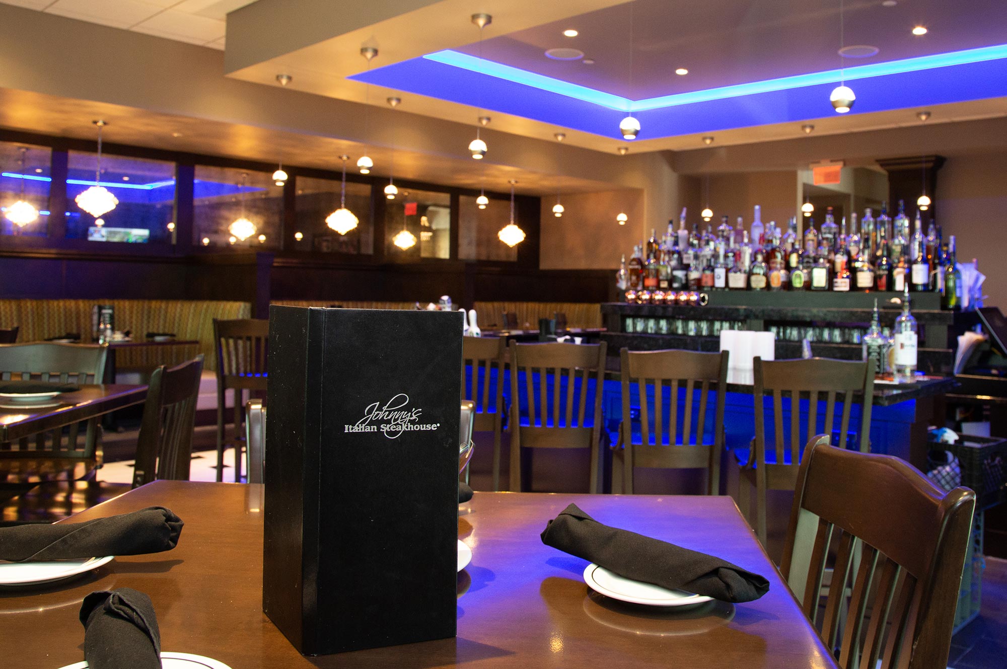 A modern restaurant interior featuring wooden tables set with black napkins, a menu, and a bar area in the background stocked with various bottles under blue lighting.