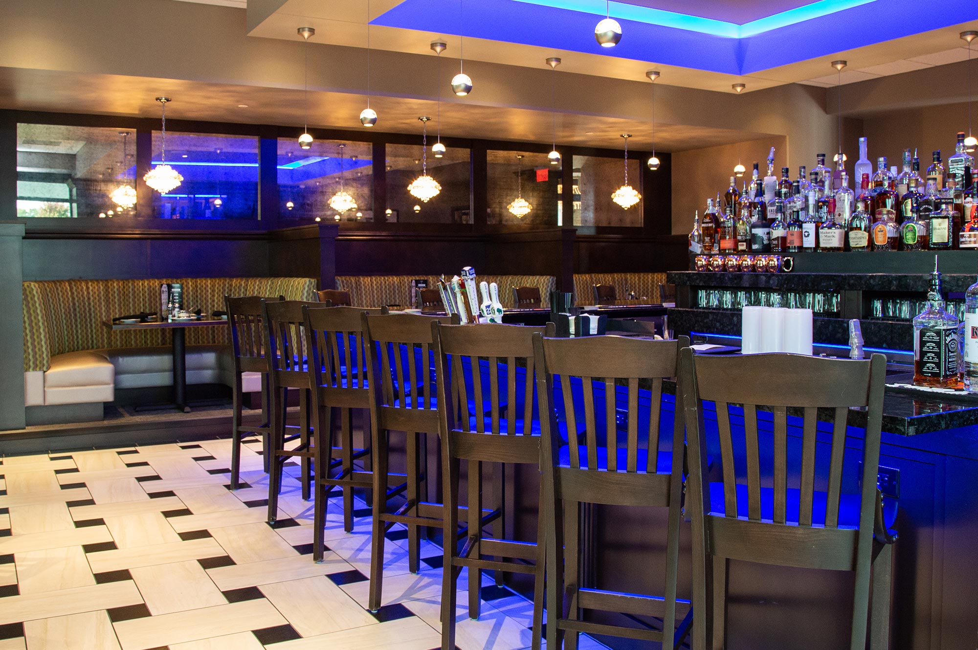 A modern bar with wooden high chairs, a backlit counter, hanging pendant lights, and various bottles of liquor on shelves. Booth seating is visible in the background.