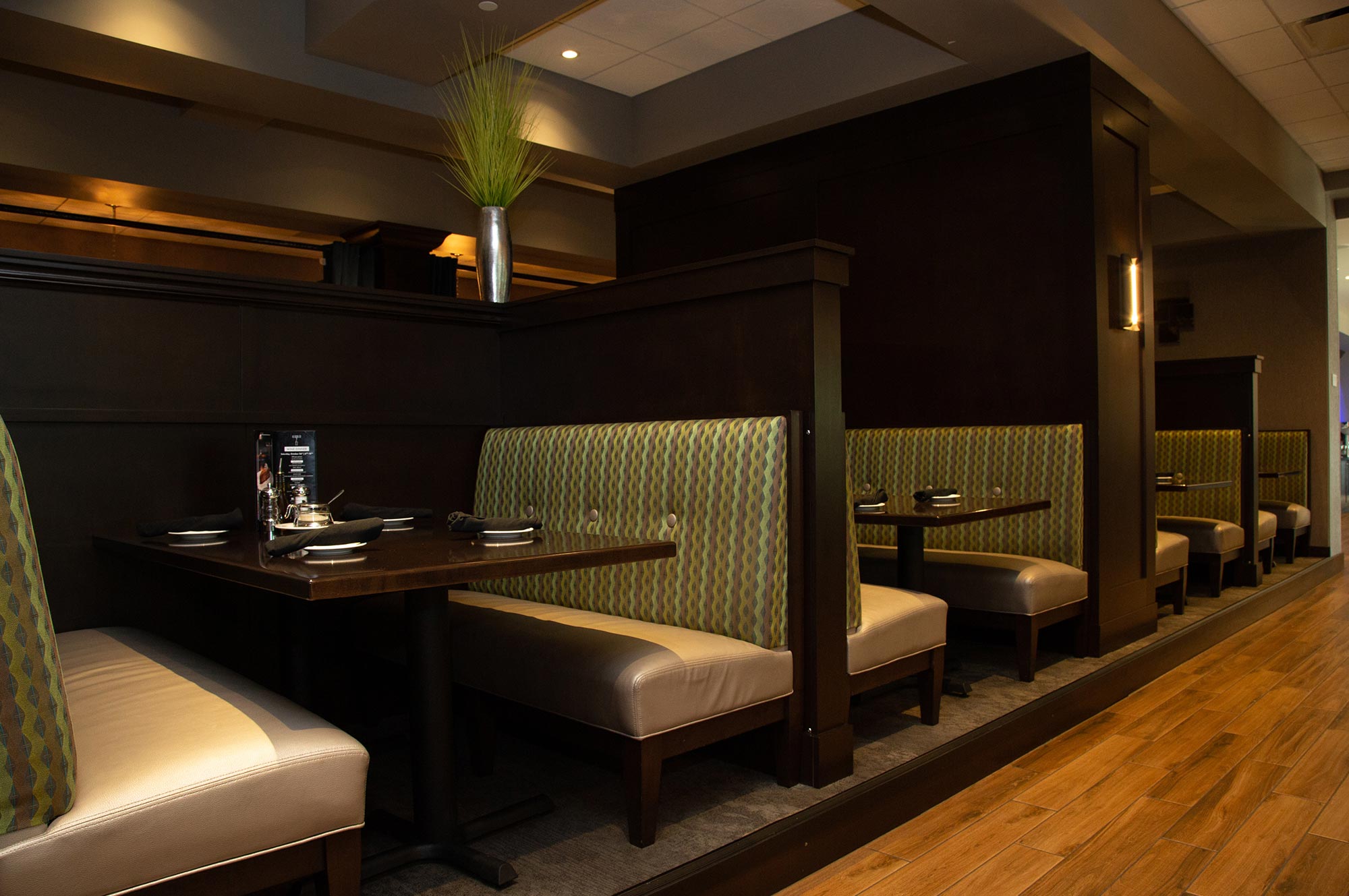 Interior of a restaurant featuring several empty booths with patterned upholstery, neatly set tables with plates and cutlery, and wooden flooring.