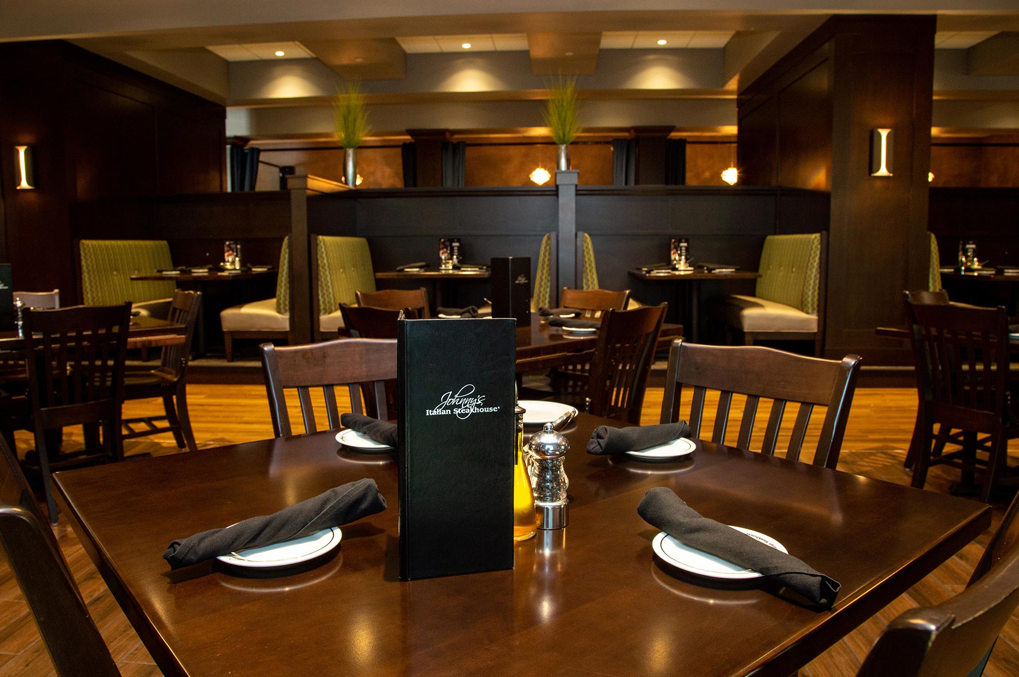 A dimly lit restaurant interior featuring wooden dining tables set with black napkins, white plates, and a menu at the center. Booth seating is visible in the background.