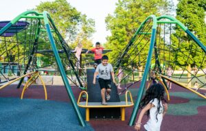 Three children play on a jungle gym in an outdoor playground on a sunny day, surrounded by trees and green grass.