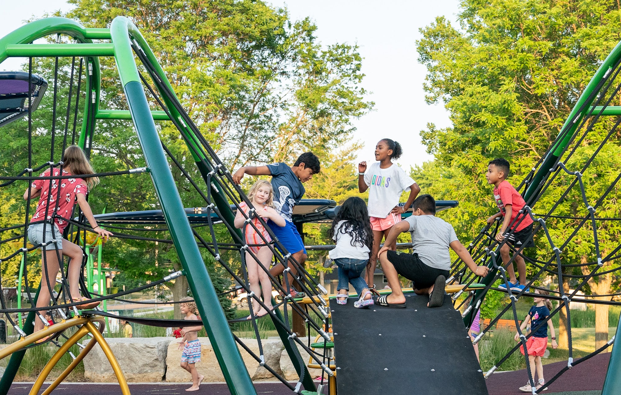 Children are playing and climbing on a jungle gym in a playground surrounded by trees on a sunny day.