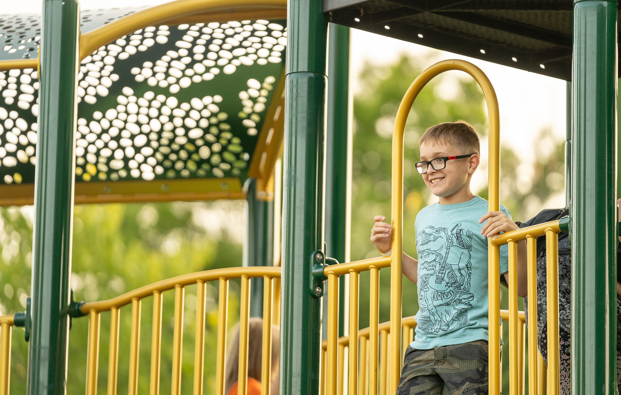 A boy wearing glasses and a blue shirt stands smiling on a play structure with yellow railings at a playground.