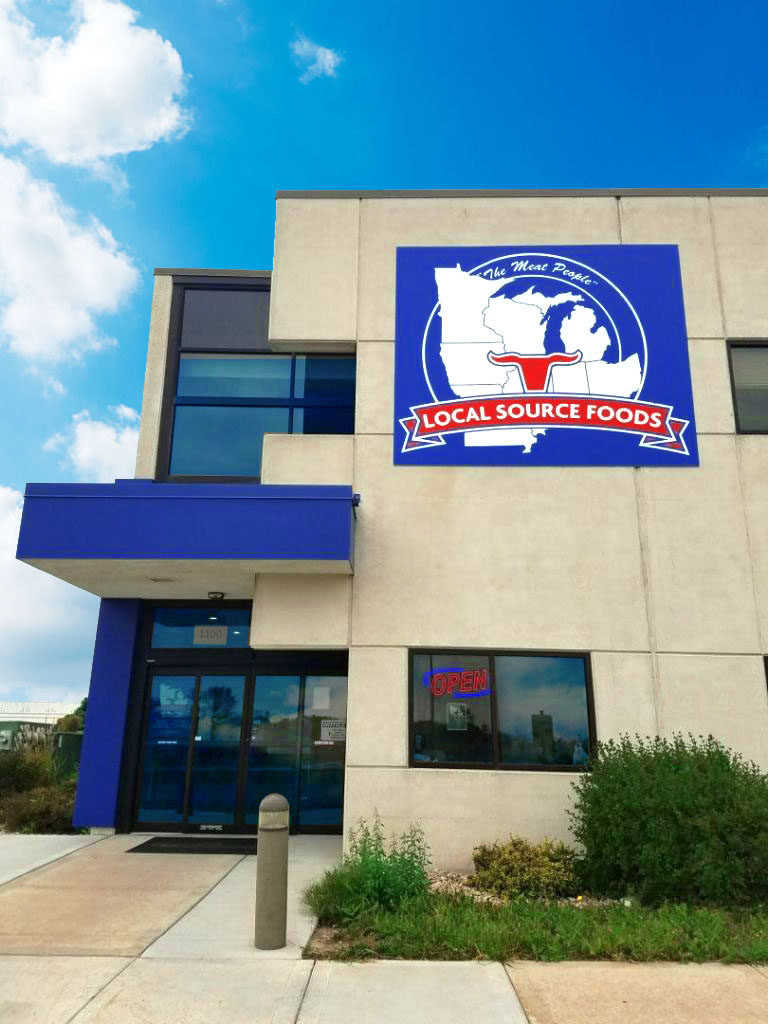 A beige and blue building houses Local Source Foods. A large logo depicting the state of Michigan is displayed on the wall. The entrance has glass doors, one marked "Open." A clear sky is visible.