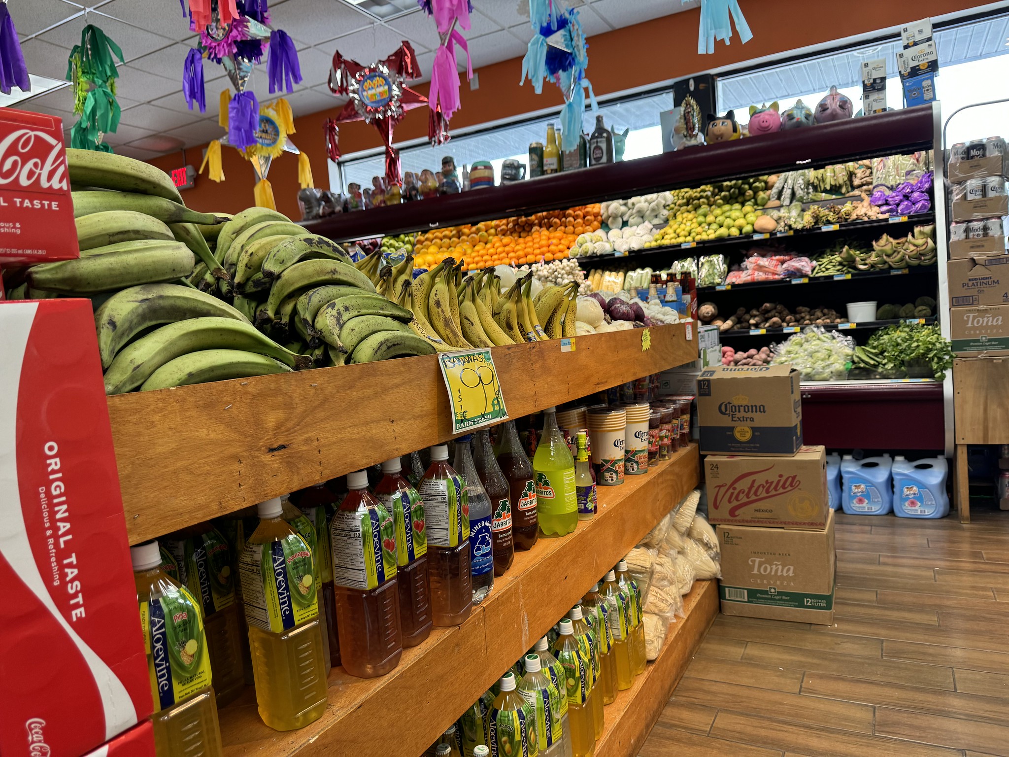 A grocery store aisle with various fruits and vegetables displayed on shelves. Bottles of beverages are stacked below the produce. Colorful decorations hang from the ceiling.