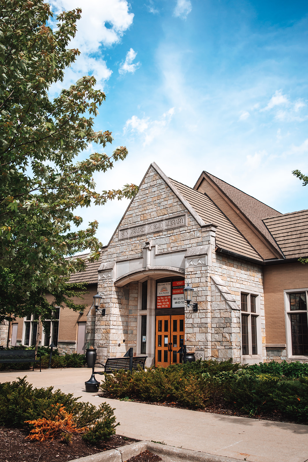 An exterior view of a stone public library building with a pointed roof under a partly cloudy sky, surrounded by trees and shrubs.