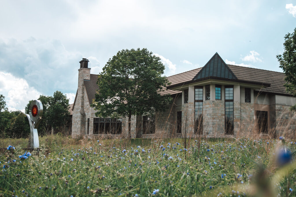 A stone building with a pitched roof stands behind a field of tall grasses and wildflowers. A tree is near the center, and a sculpture of a person holding a red circular object is on the left.