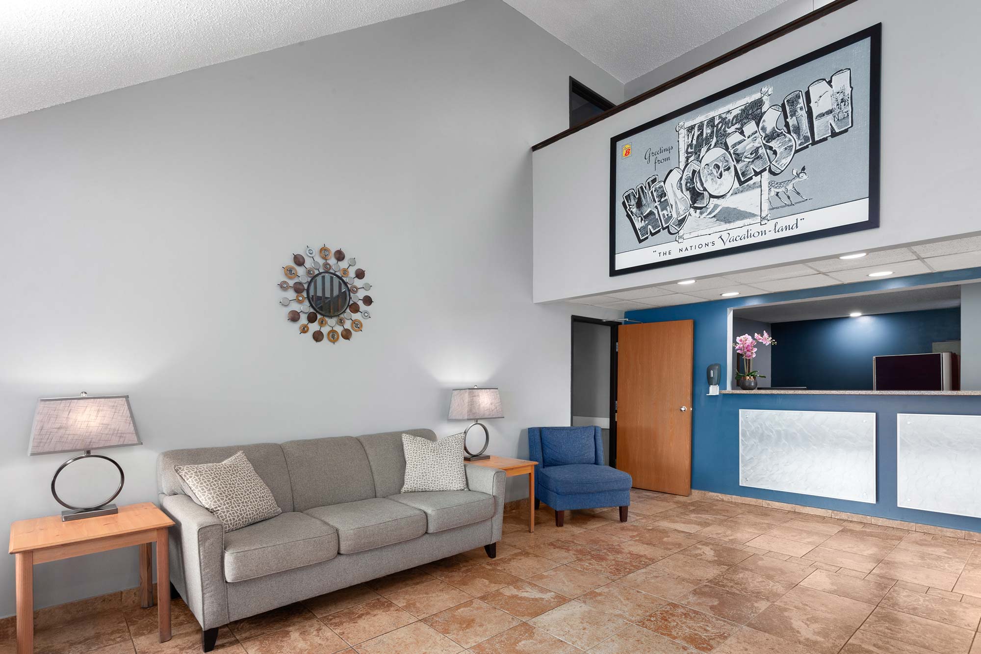 Hotel lobby with a gray couch, blue chair, and tan tile flooring. A round mirror and two lamps are on the wall, and a large framed poster hangs above. The reception desk is seen to the right.