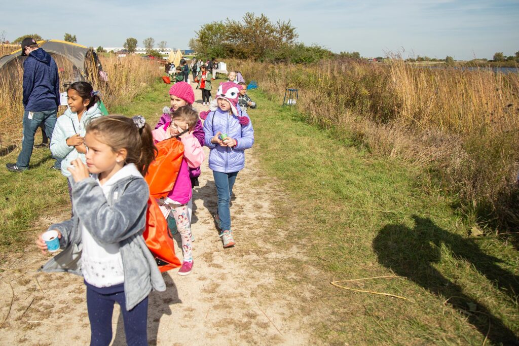 Children and adults walk along a grassy path near tall reeds and tents on a sunny day. Some children are wearing costumes, likely for a festive event.