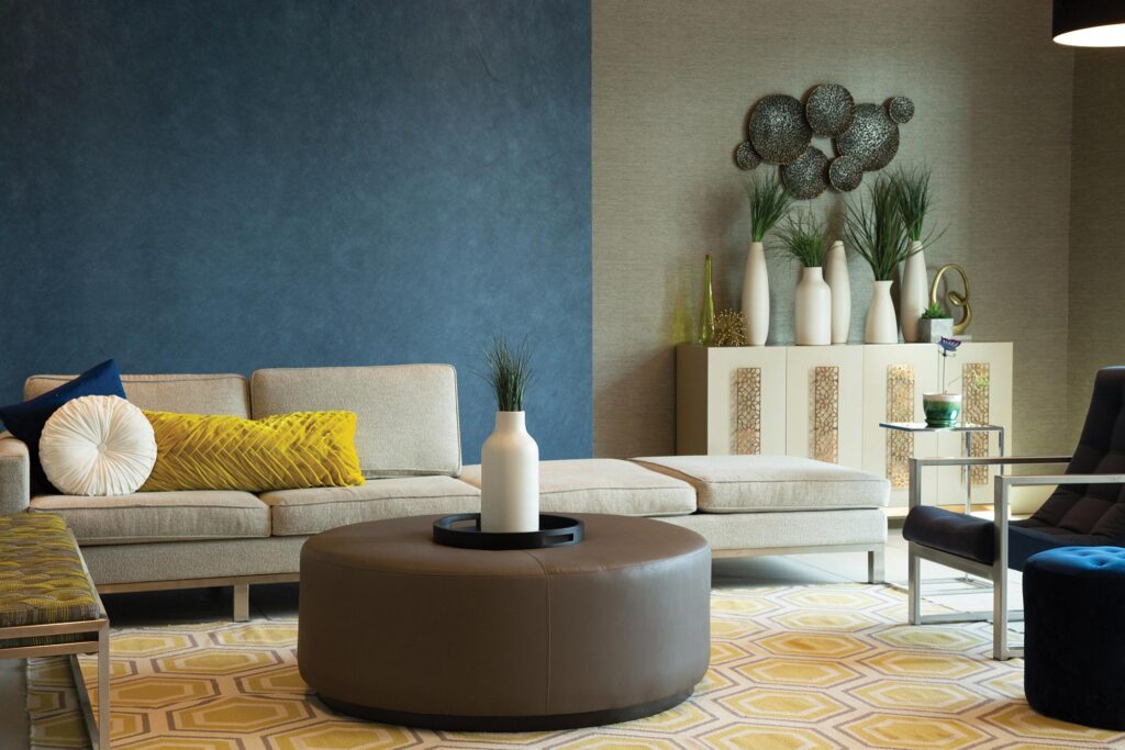 A modern living room featuring a beige sectional sofa with yellow and white pillows, a round brown ottoman, a white vase centerpiece, geometric wall art, and a blue accent wall.