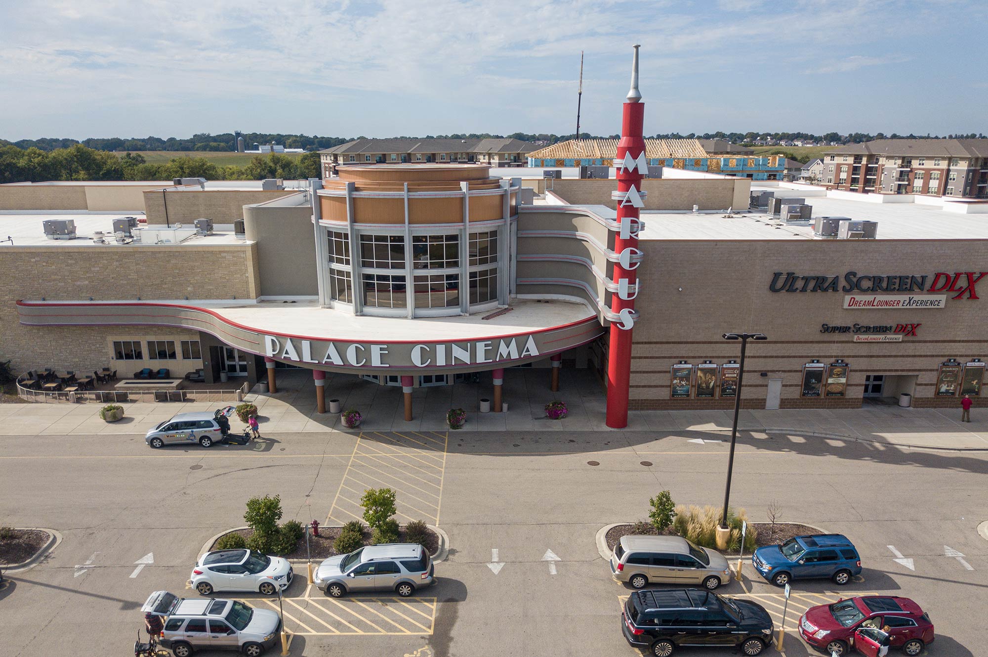 Aerial view of a large cinema complex with a "Palace Cinema" sign, multiple movie posters, and a parking lot with several cars. The surrounding area includes trees and other buildings.