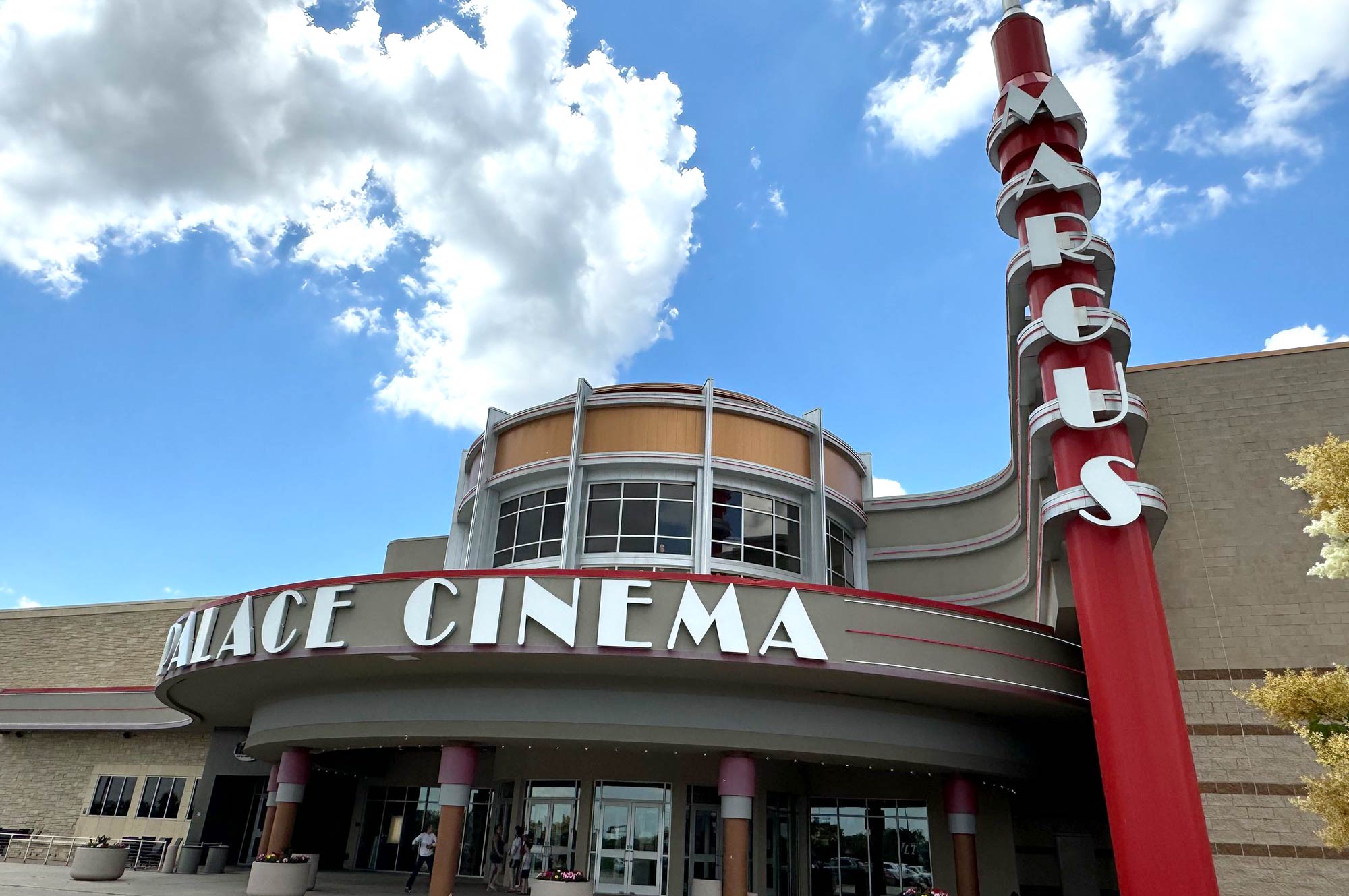 A large movie theater with a sign reading "Palace Cinema" and a tall vertical sign displaying "Marcus". The building features a rounded entrance with glass windows and a red column.