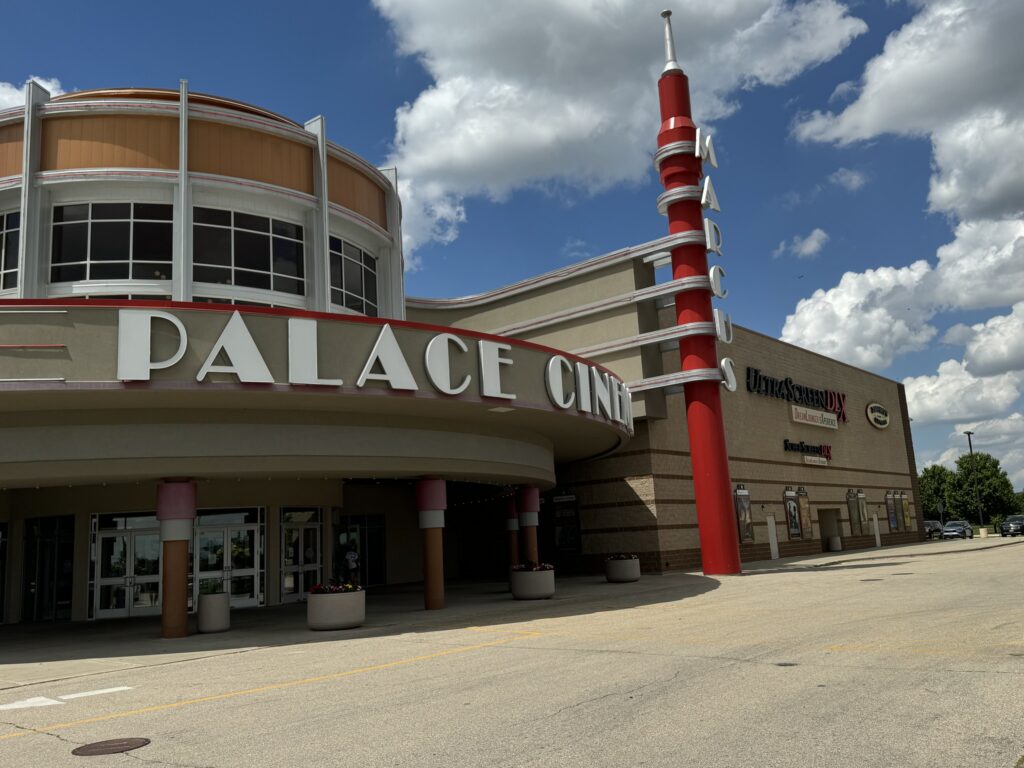 Exterior of Marcus Palace Cinema with a rounded entrance and distinctive red vertical sign under a partly cloudy sky. Several entry doors and a building extension can be seen.