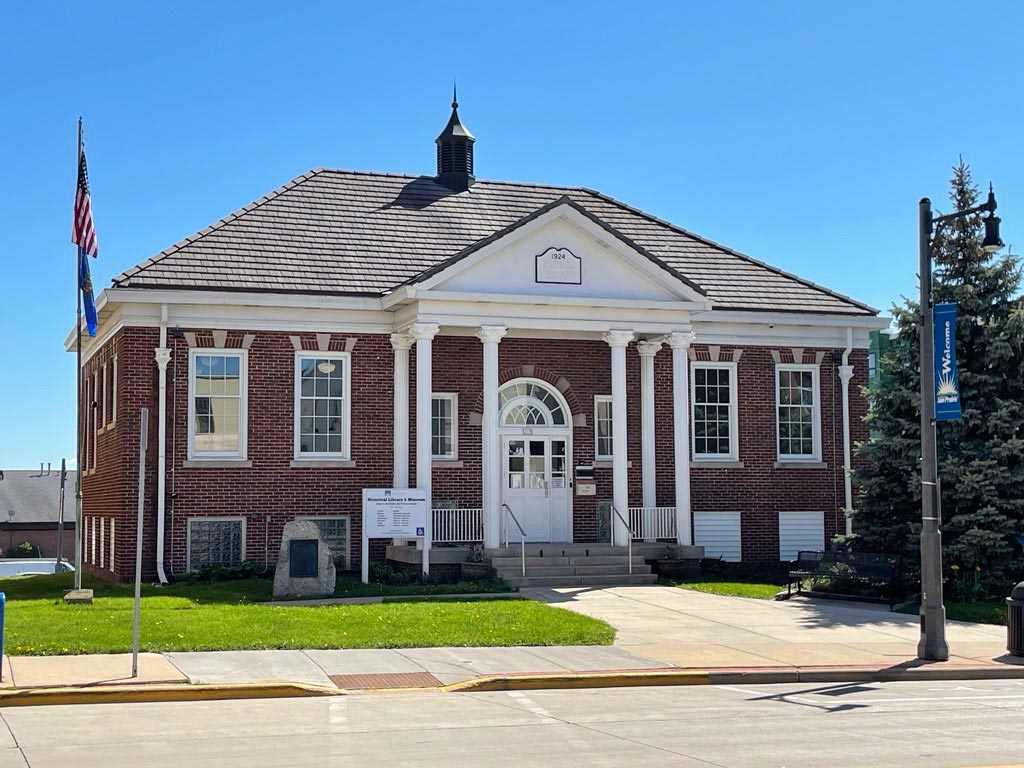 A red brick, single-story municipal building with white columns, a peaked roof, and American flags prominently displayed outside. The surrounding area includes a sidewalk, trees, and a streetlamp.