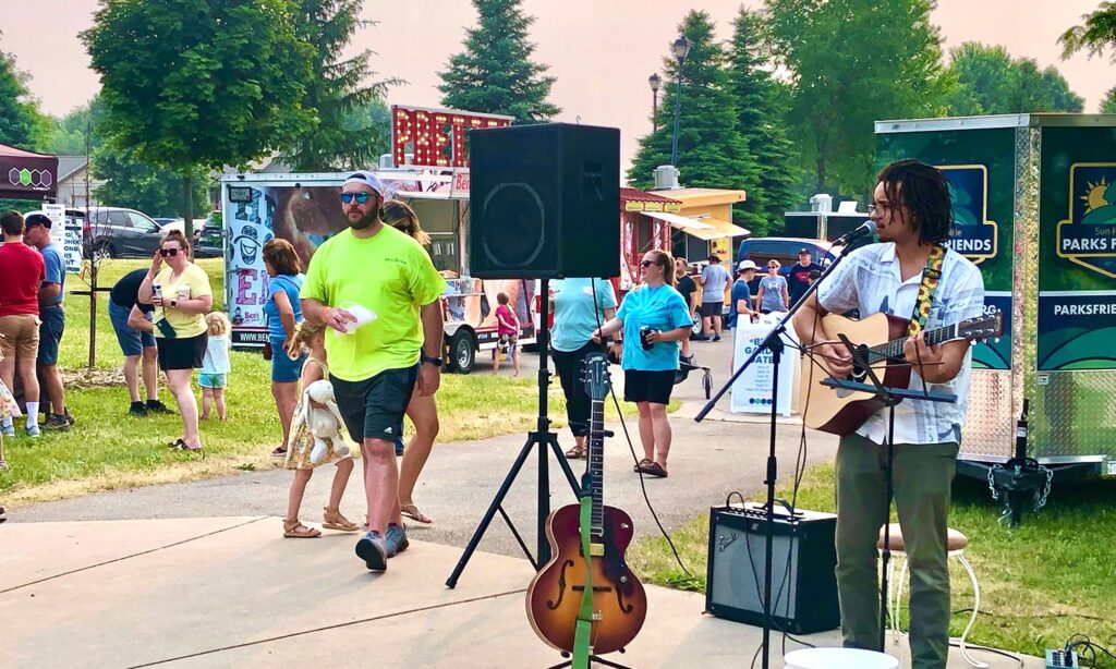 A street performer plays the guitar and sings near various food trucks, while people walk and gather around in a park setting on a sunny day.