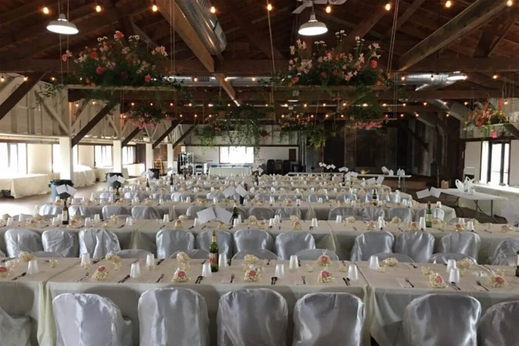 A large rustic hall is set up for an event with rows of tables and white chairs, floral centerpieces, and hanging flower decorations under a wooden ceiling with string lights.