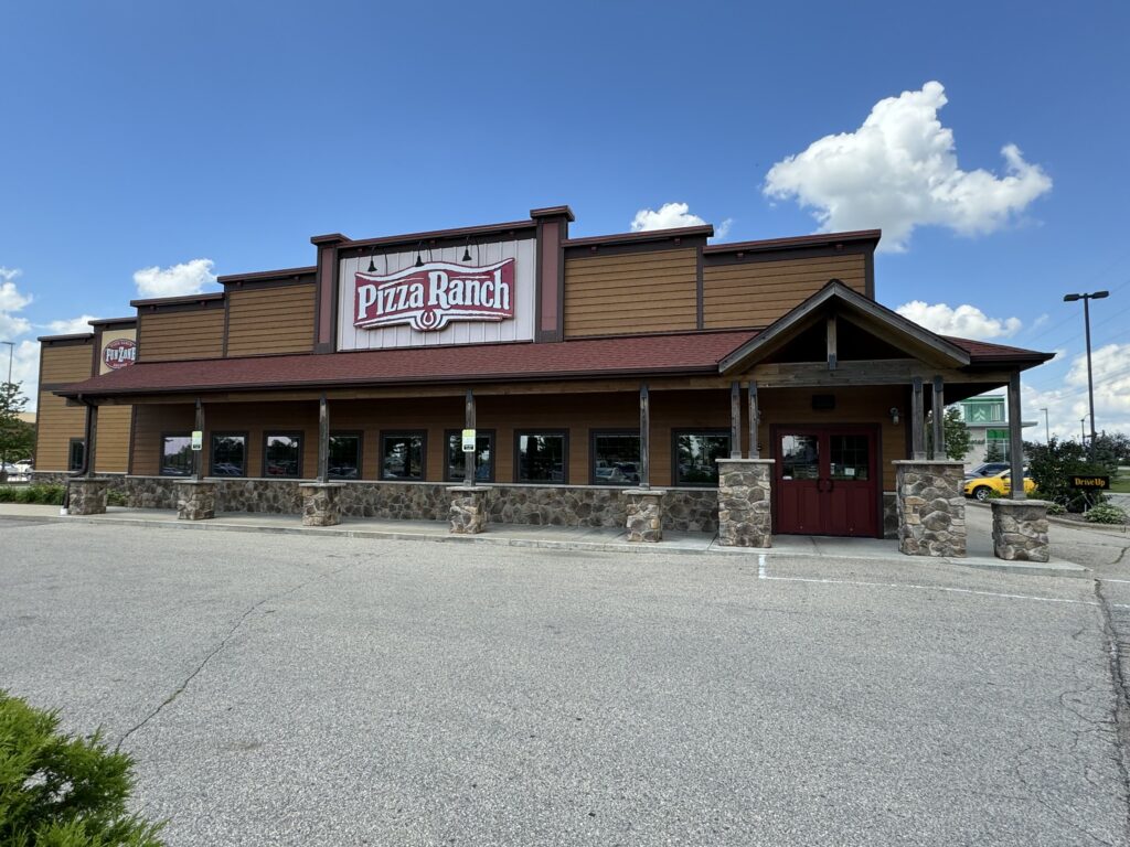 A Pizza Ranch restaurant with a tan and brown exterior, stone accents, and a red door. The parking lot in front is nearly empty under a partly cloudy sky.