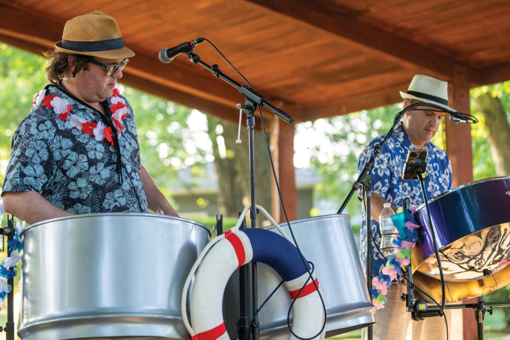 Two musicians perform on steel drums under a wooden pavilion. Both are wearing floral shirts and hats. One microphone is positioned between them, and a life preserver is attached to a drum.