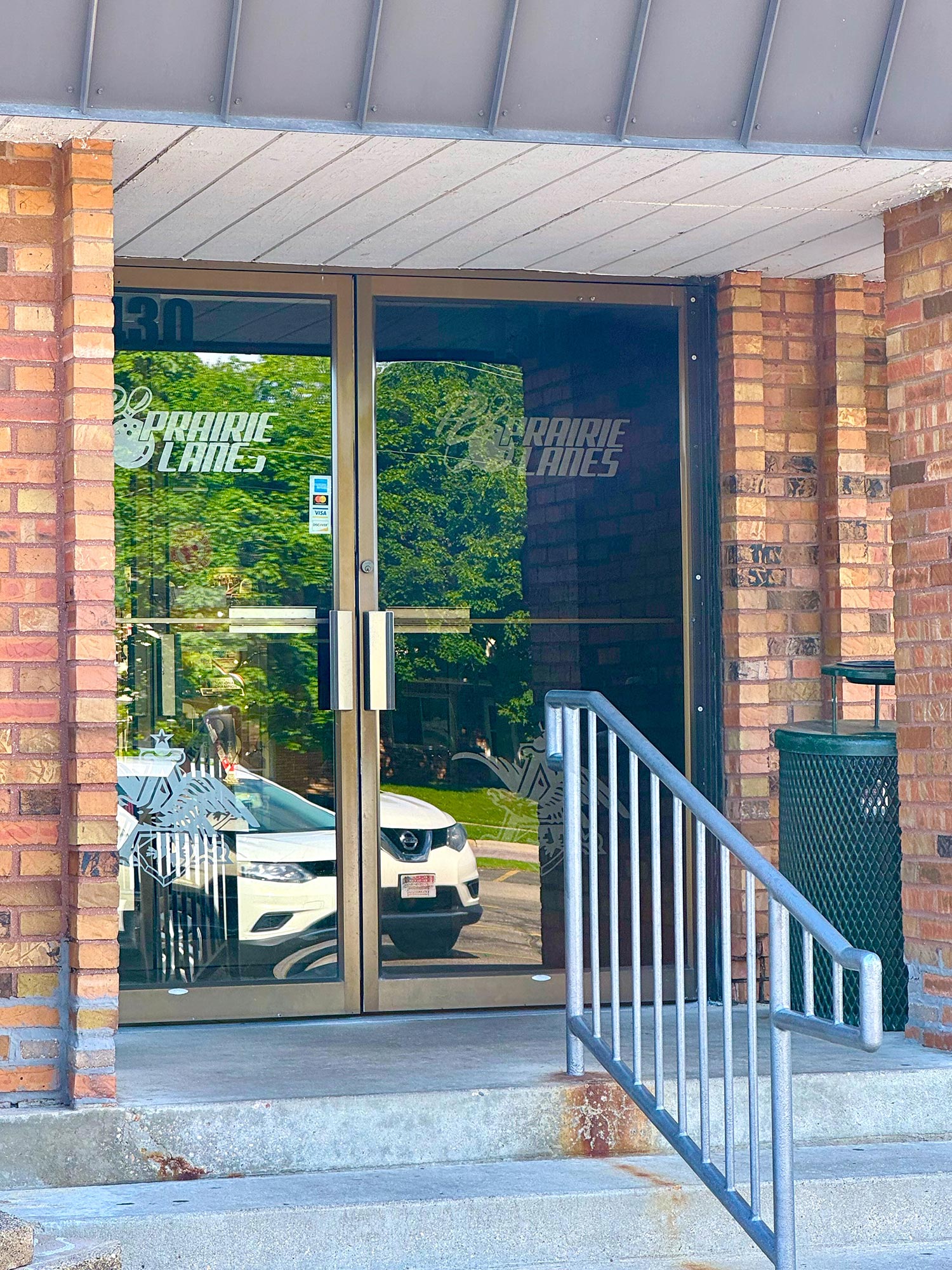 Glass entrance door of a brick building, reflecting a parked white car and trees. "Prairie Lanes" is on the door. Metal railing leads to the door with a dark green trash can to the right.