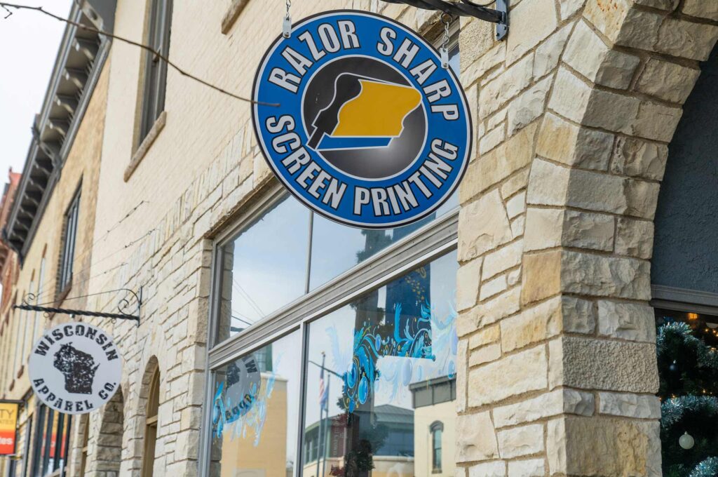 Storefront of Razor Sharp Screen Printing with a blue and yellow logo, next to Wisconsin Apparel Co. signs and stone archways.