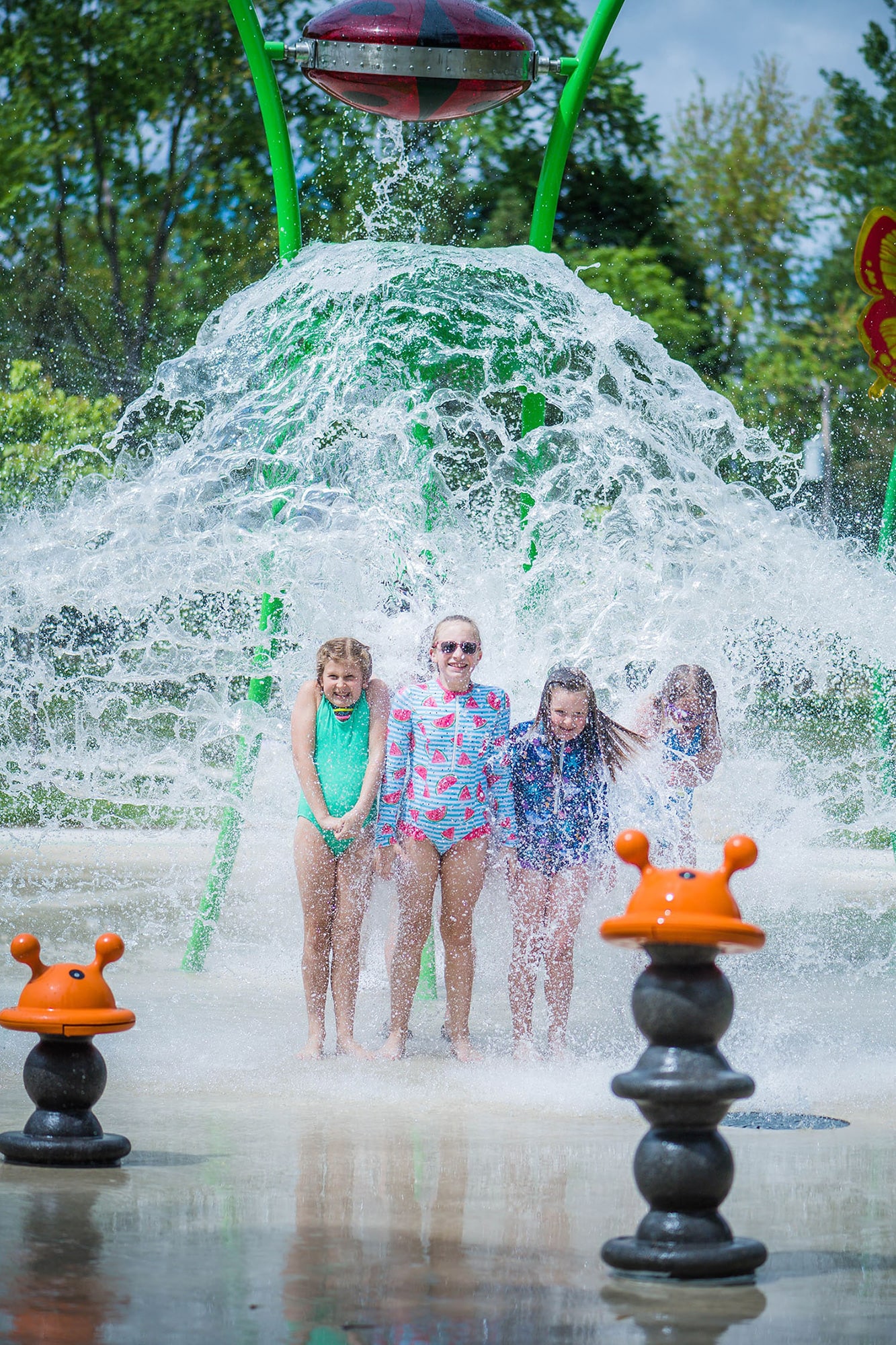 Four children stand underneath a large water feature at a splash park, getting soaked by the water cascading down.