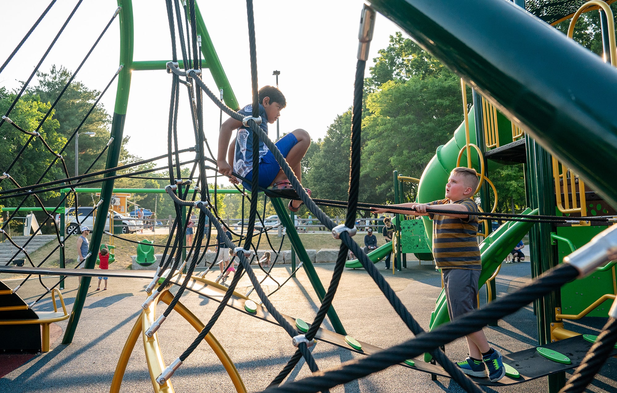 Two children play on a jungle gym made of ropes and metal bars at a playground. One child is climbing while the other is standing on the ground holding onto the structure. Trees and other playground equipment visible.