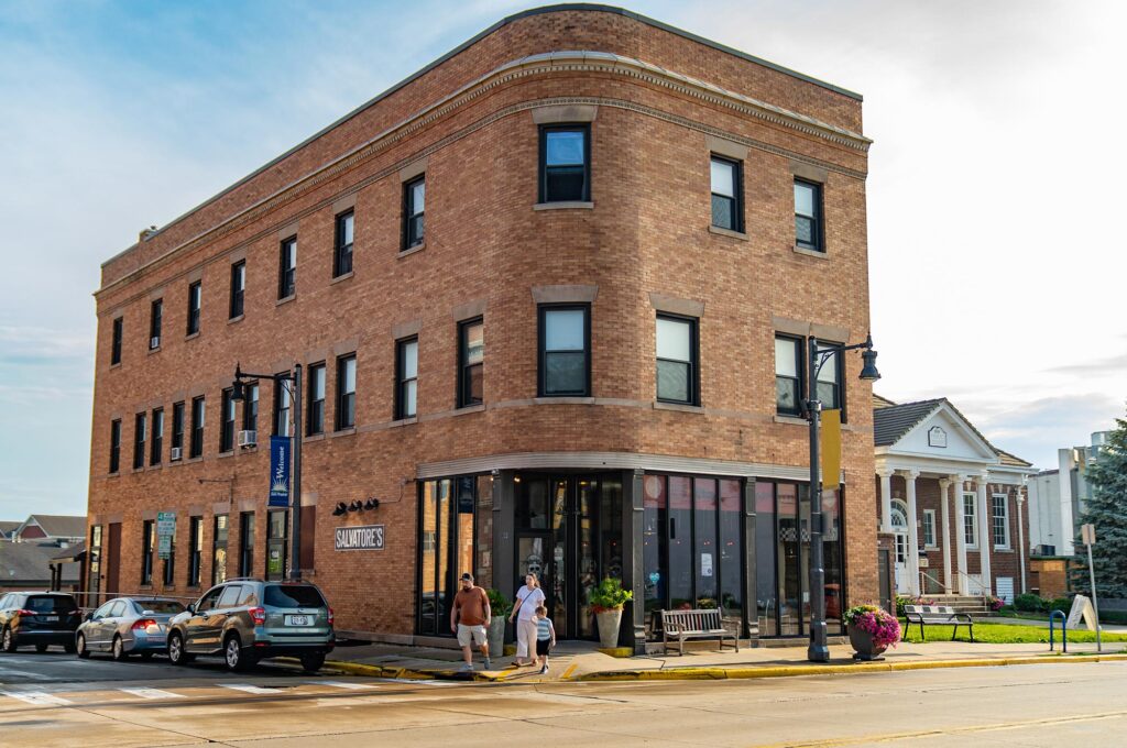 A three-story brick building with large windows on the corner of a street. Two people are walking towards the entrance, and several cars are parked along the street.