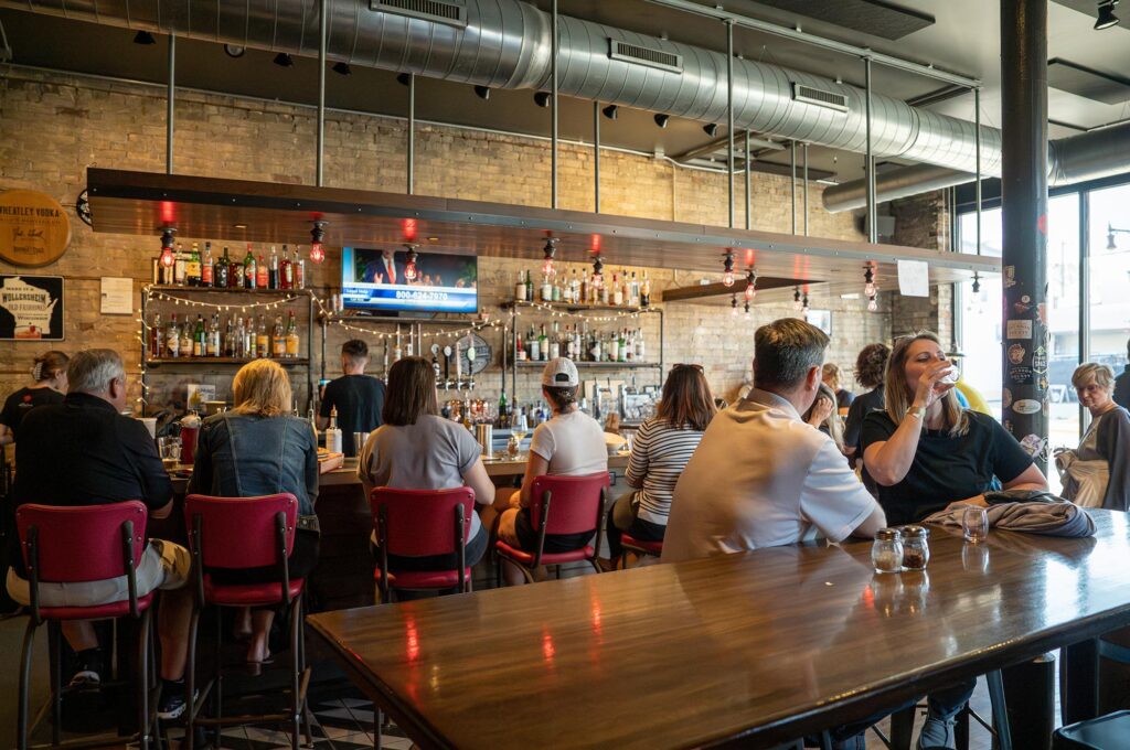 People sitting at a bar in a restaurant with a TV mounted on the wall above the bar. Various bottles of alcohol are displayed on shelves behind the bar. Daylight is coming through the windows.