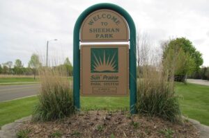 Sign reading "Welcome to Sheehan Park" with "City of Sun Prairie Park System" logo, surrounded by tall grass, near a road with trees in the background.