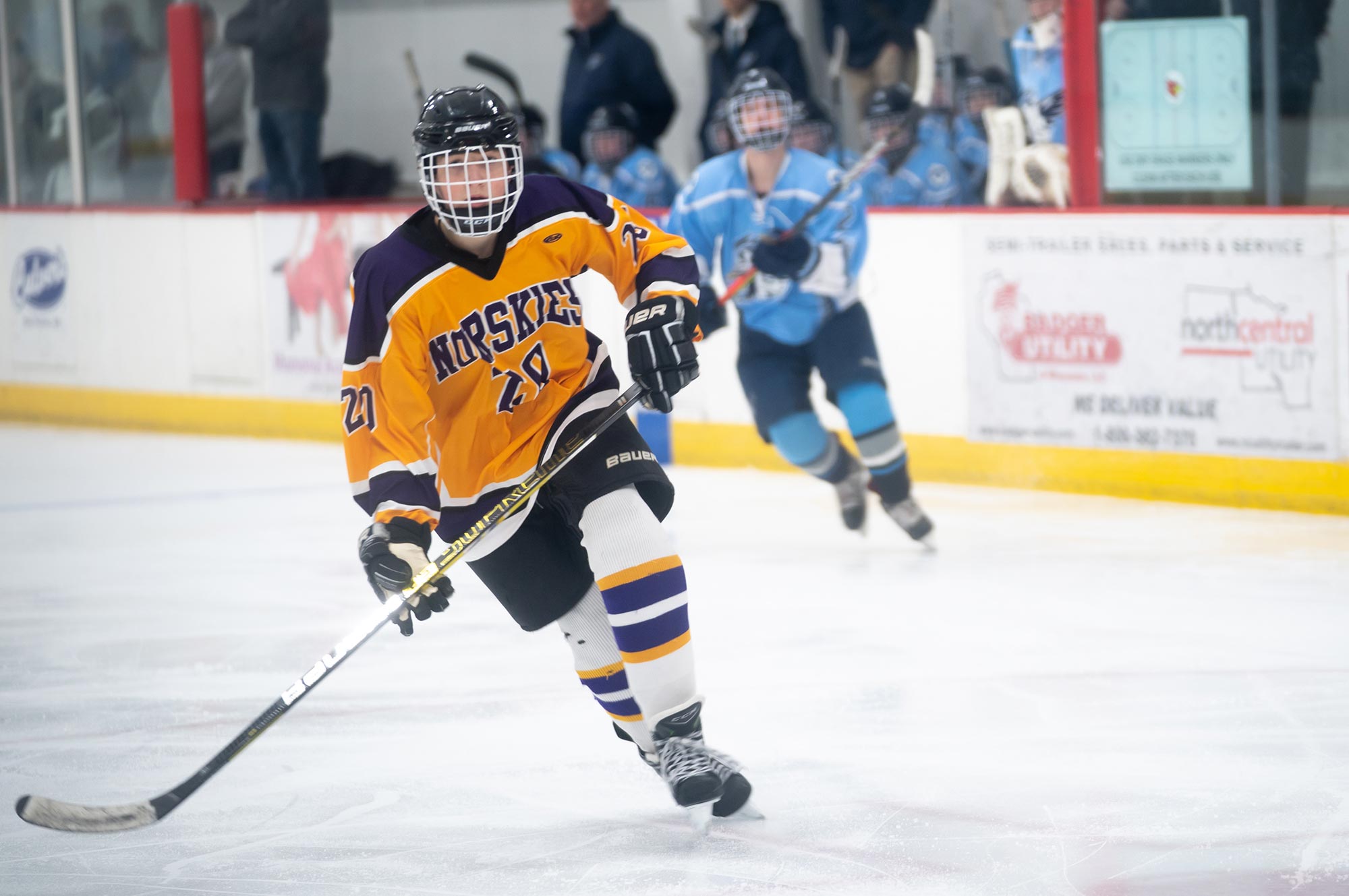 A hockey player in a yellow and purple uniform skates with the puck while another player in light blue pursues from behind on an indoor ice rink.