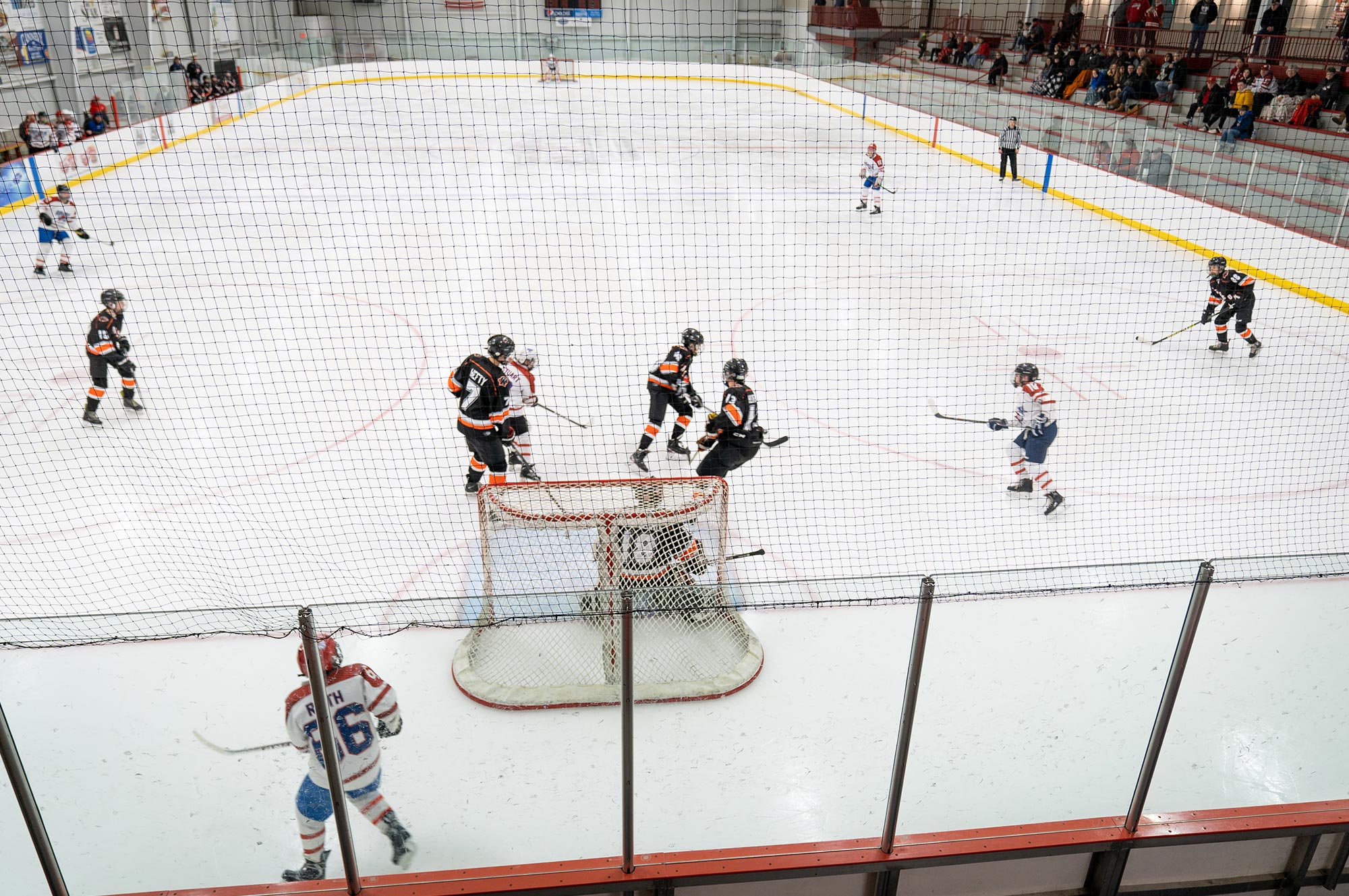 Ice hockey game in progress, players in red and white facing players in black and orange. A player attempts a shot at the goal with the goalie ready to block.