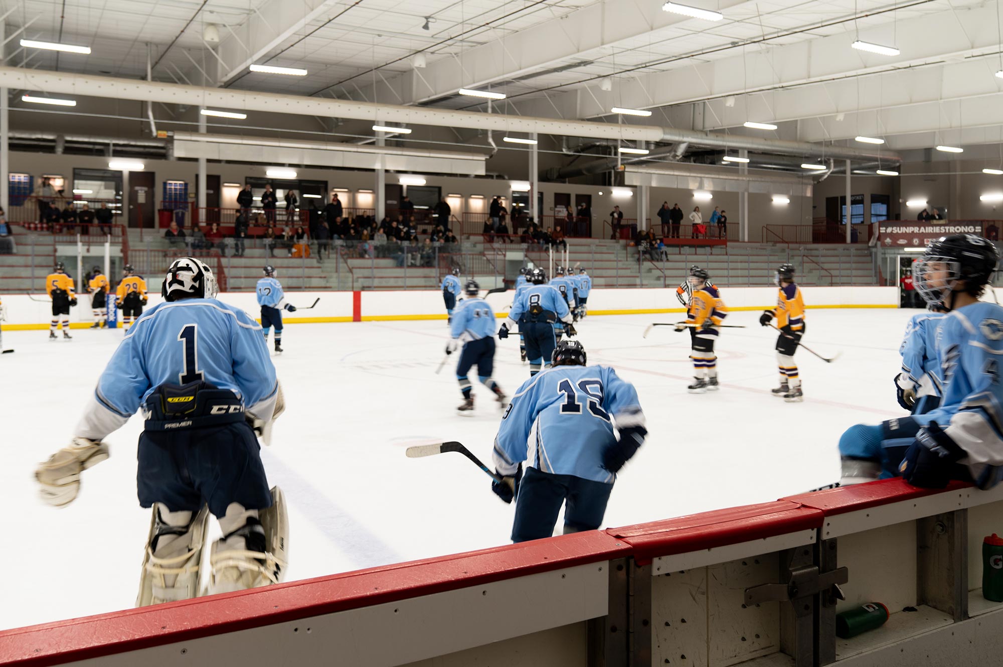 Youth ice hockey game in progress at an indoor rink. Players in light blue jerseys are on the ice and bench, while players in orange jerseys face them. Spectators are seated in the stands.