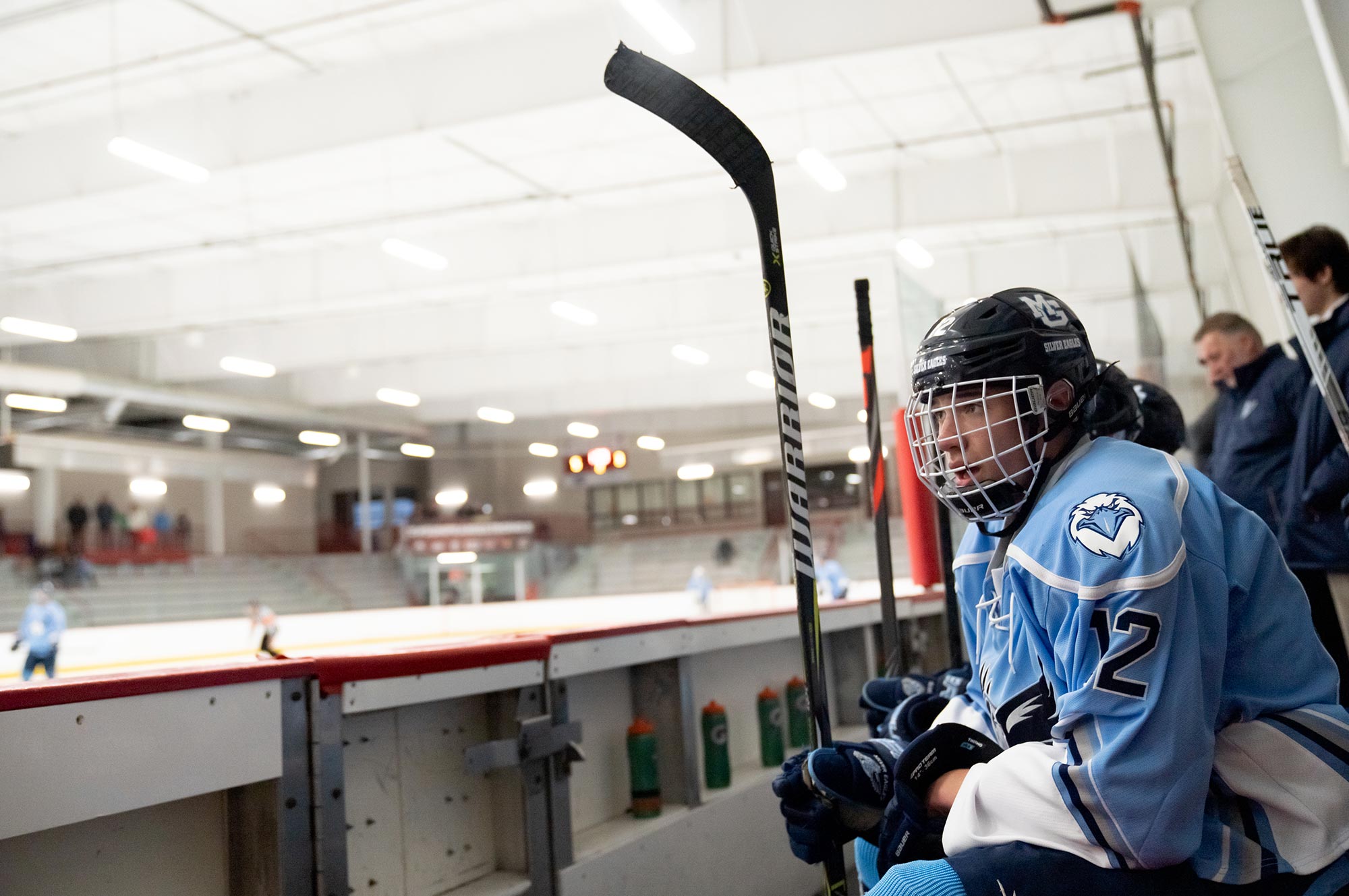 A hockey player dressed in a blue and white uniform sits on the bench inside an ice rink, holding a hockey stick, with other players and a coach behind him.