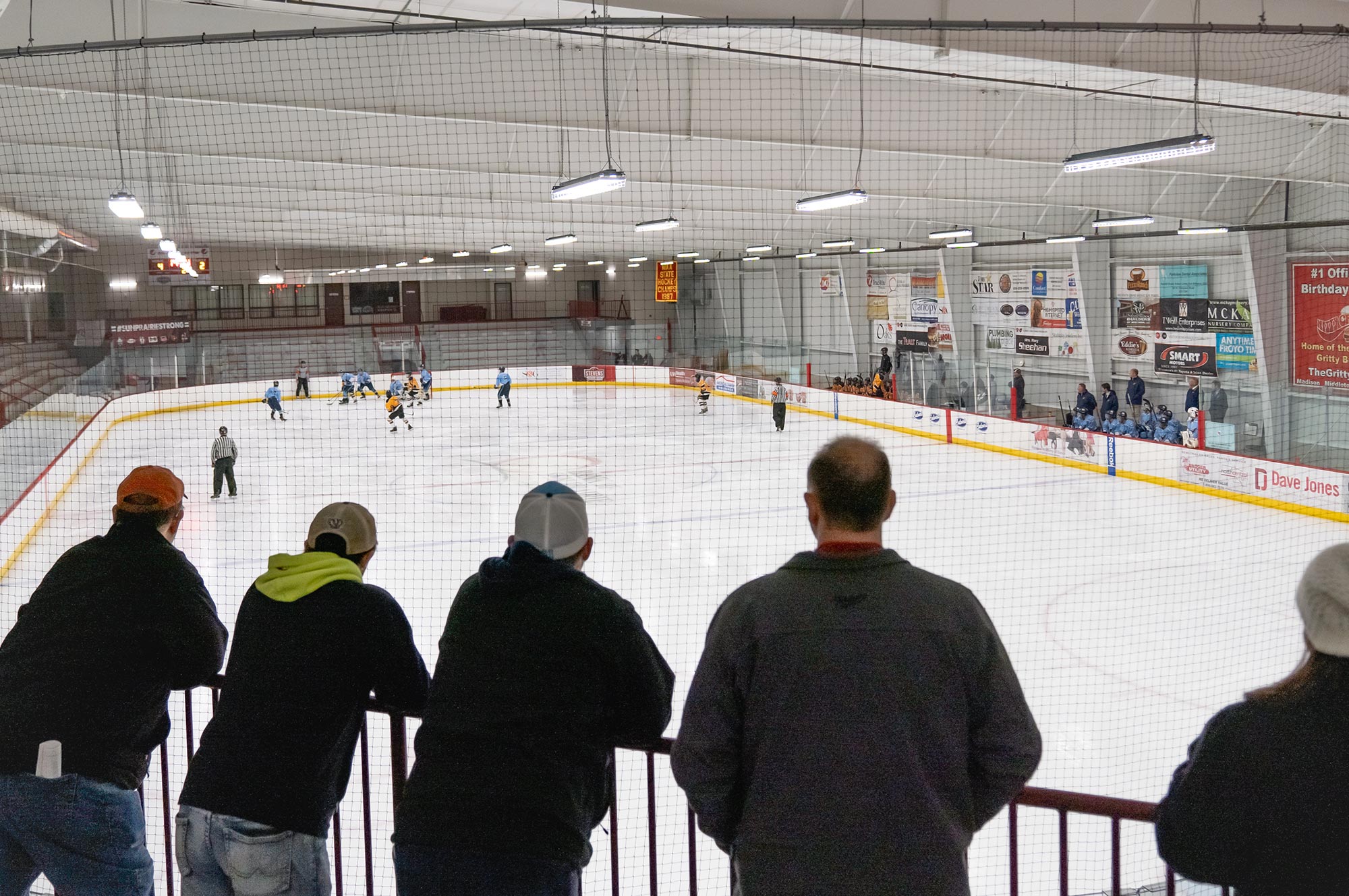 A group of people watches a hockey game at an indoor ice rink from behind a protective net. Players in blue and yellow jerseys are on the ice, while coaches and teammates observe from the benches.