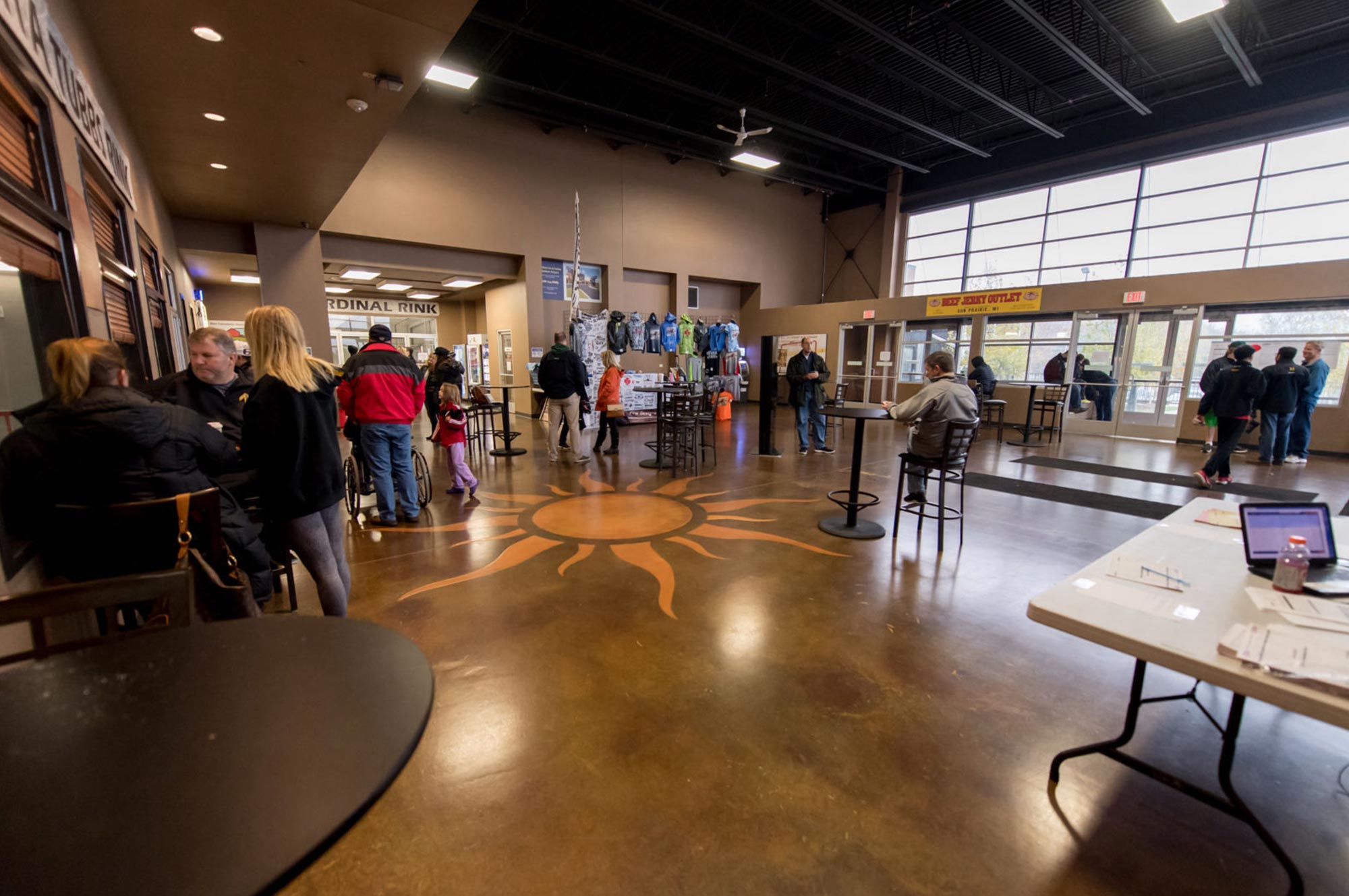 People are gathered inside a lobby with large windows and tables. Some are seated, others are standing in line. The floor has a sun design. A countertop with a laptop is visible.