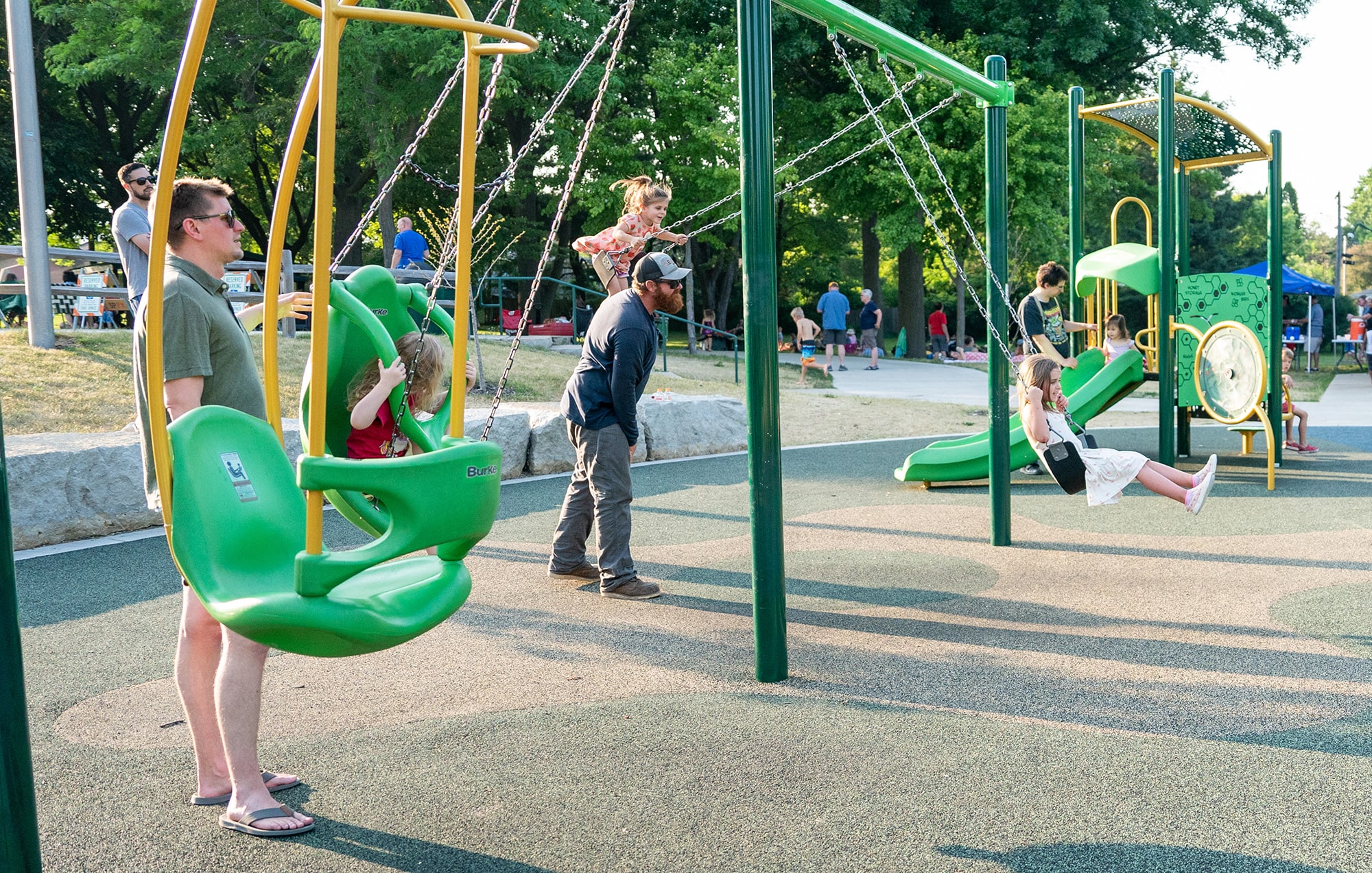 People enjoying a playground; two adults pushing children on swings, another child on slide. Green play equipment and trees in background. Sunny day with some people in the distance.