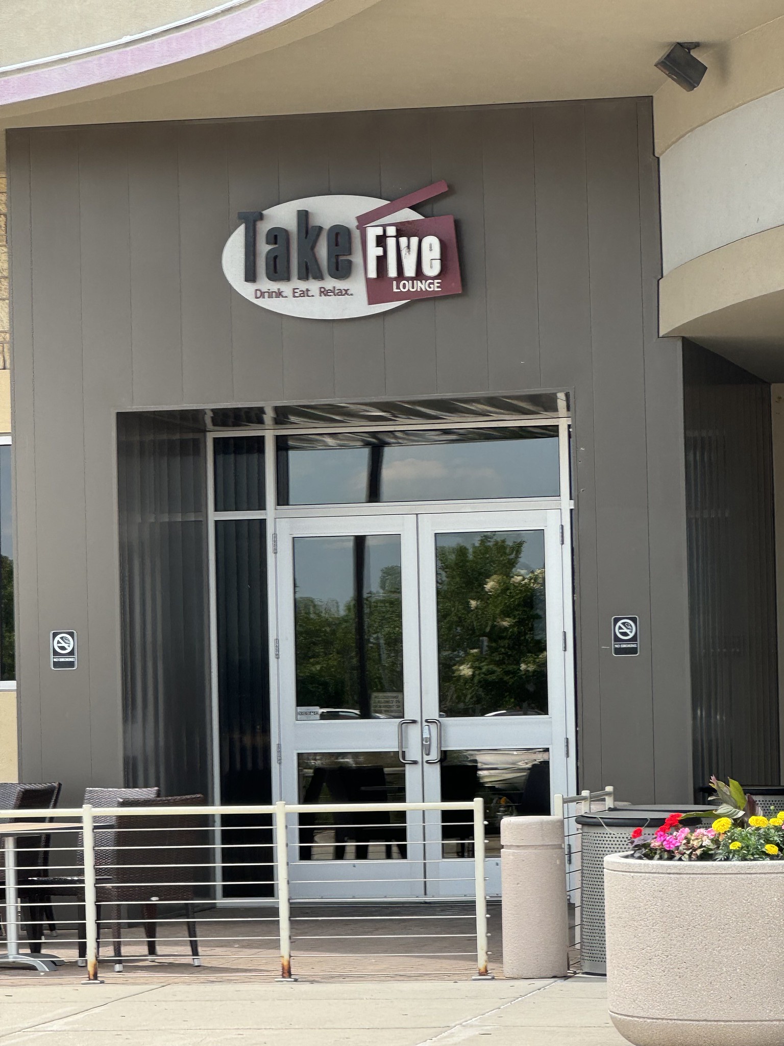 Exterior view of a lounge called "Take Five" with a sign reading "Drink. Eat. Relax." There are tables and chairs outside, and flower pots near the entrance.