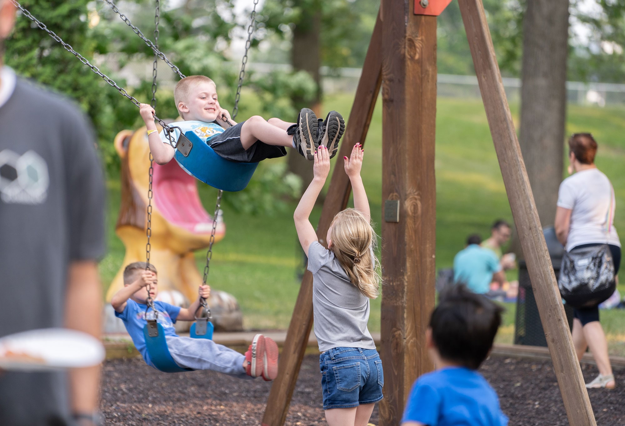 Children playing on swings at a park, with a girl pushing a boy on a swing while other kids and adults are visible in the background.