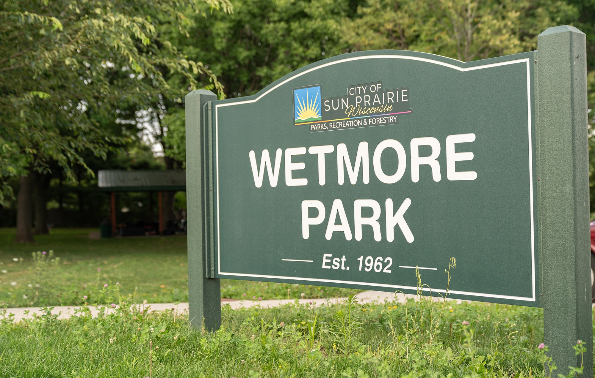 A green sign with white letters reads "Wetmore Park, Est. 1962" in Sun Prairie, Wisconsin. Trees and a shelter are visible in the background.