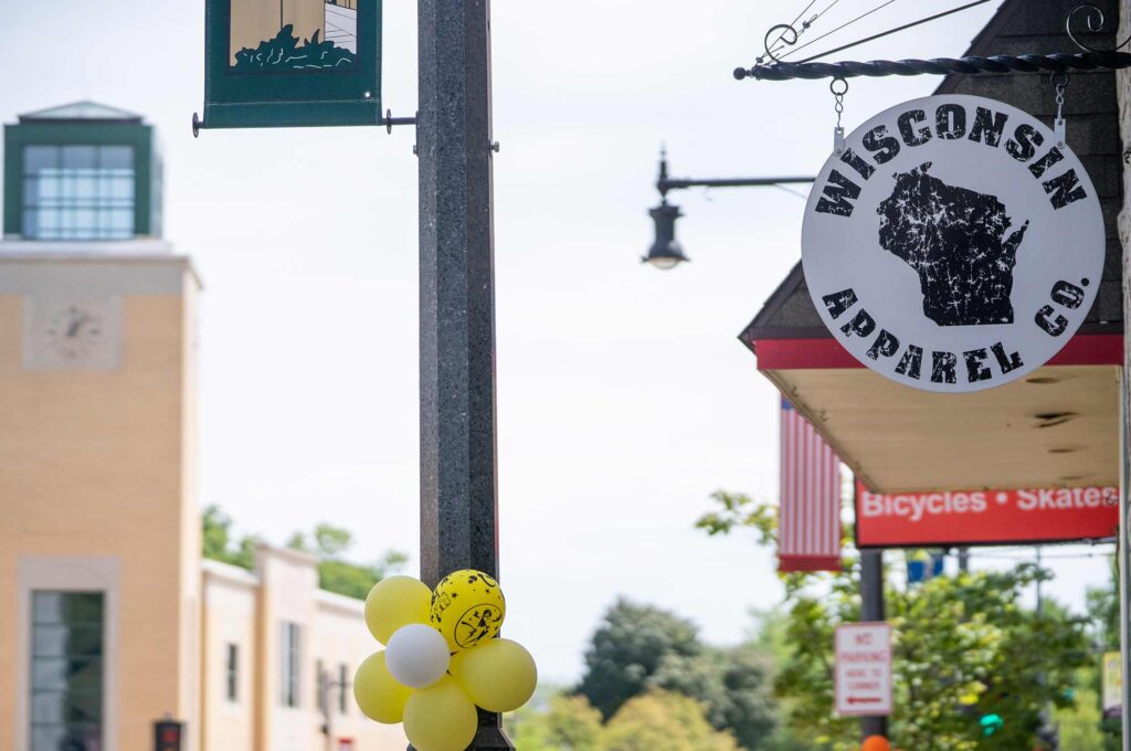 A street scene features a store sign reading "Wisconsin Apparel Co." with a state outline, next to a building with a clock. Yellow balloons are attached to a lamppost.