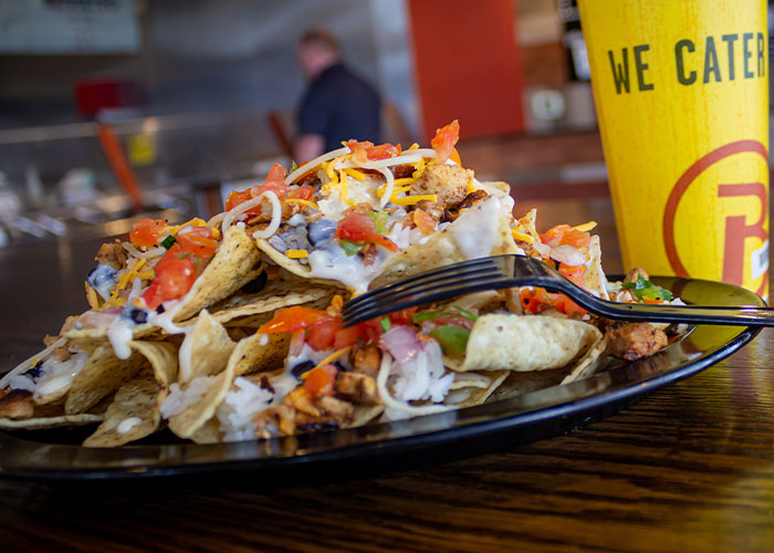 A plate of loaded nachos topped with tomatoes, shredded cheese, sour cream, and green onions next to a yellow beverage cup on a wooden table in a restaurant setting.