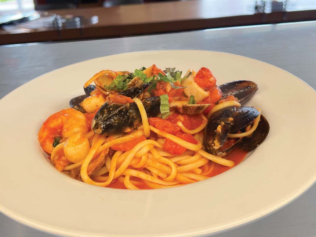 A white plate of linguine with seafood including shrimp, mussels, and clams in a tomato-based sauce, garnished with chopped herbs.