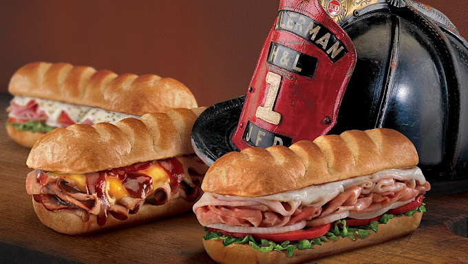 Three assorted sub sandwiches with meat and vegetables are placed on a wooden surface next to a firefighter's helmet with an insignia labeled "UFD 1.