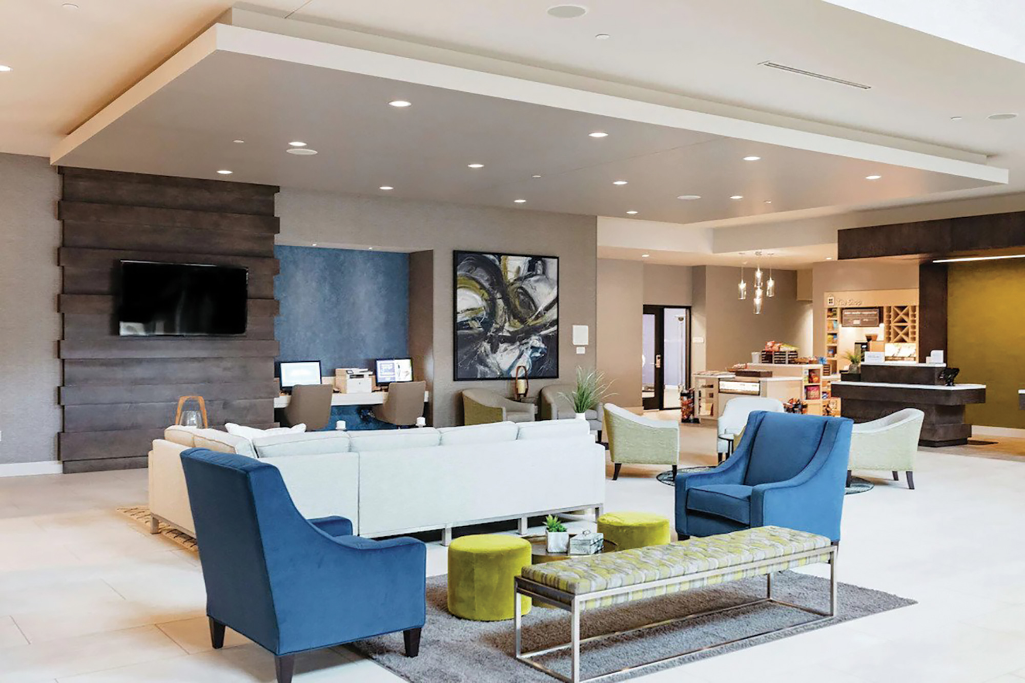 A modern hotel lobby with blue and green chairs, a white sofa, a wall-mounted TV, a reception desk, and a painting on the wall. The area is well-lit with ceiling lights.