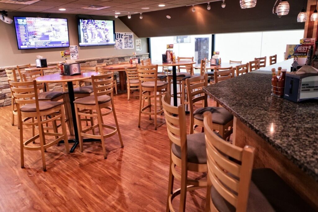 A bar interior with wooden chairs and tables, a granite countertop, and multiple TV screens showing sports. The bar is well-lit with pendant lights hanging from the ceiling.